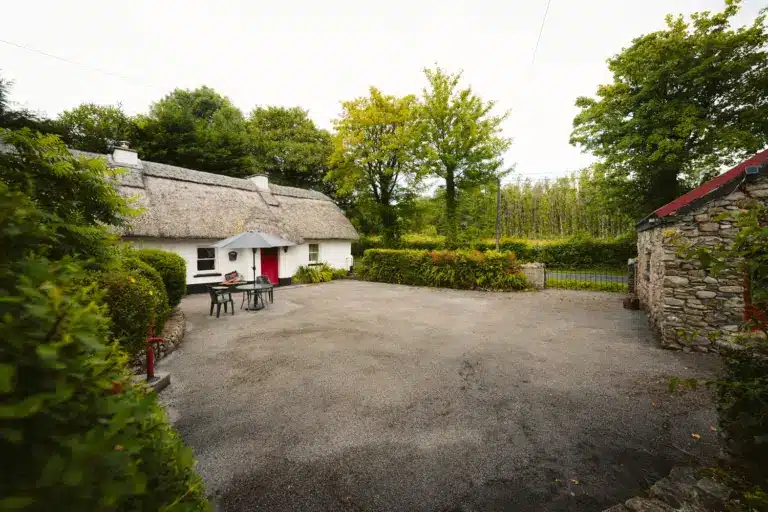 A quaint white cottage with a thatched roof and red door is surrounded by lush greenery. An outdoor table with chairs and a parasol sits on a gravel courtyard beside a stone wall and leafy trees.