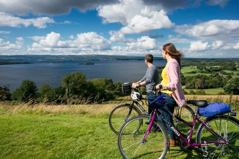 Two people with bicycles stand on a grassy hill, looking out over a large lake and a scenic landscape with green fields and trees under a partly cloudy sky.