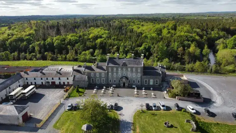 Aerial view of a large historic building with a courtyard, outdoor seating, parked cars, and surrounded by lush green trees and countryside under a partly cloudy sky.