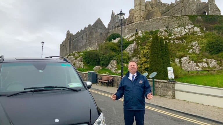 A man in a jacket stands next to a black vehicle on a street, smiling, with a historic stone castle on a hill in the background under a cloudy sky.