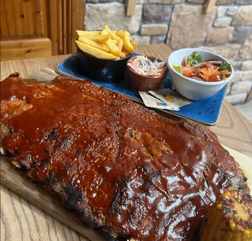 A large rack of barbecued ribs covered in sauce is served with chips, coleslaw, a bowl of salad with tomatoes, and a piece of corn on the cob on a wooden table.