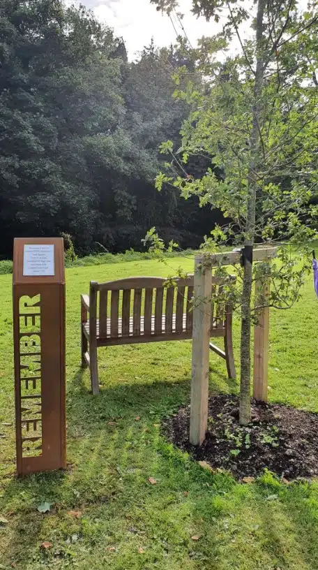 A wooden bench sits on grass beside a young tree, supported by stakes. Nearby, a metal sign reads "REMEMBER" vertically, with a plaque mounted on top. Trees and greenery fill the background.