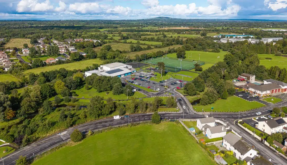 Aerial view of a suburban area with green fields, trees, sports courts, a large car park, and residential houses, all under a partly cloudy sky with distant hills in the background.