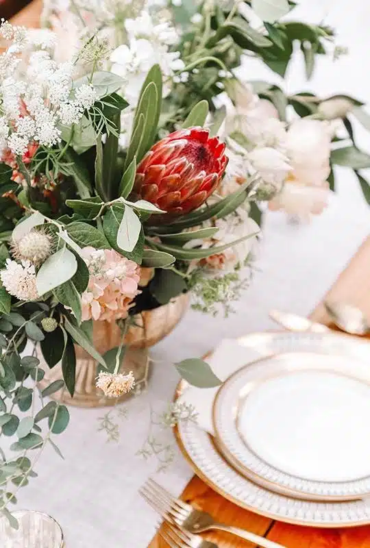A table setting with elegant gold-rimmed plates and cutlery, next to a large floral arrangement featuring greenery, white flowers, and a prominent red protea bloom in a decorative vase.