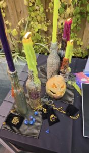 Four colourful candles in glass bottles and a carved pumpkin are on a black table with dice, small pouches, and green leafy plants in the background. Wax drips down the bottles, creating a mystical scene.
