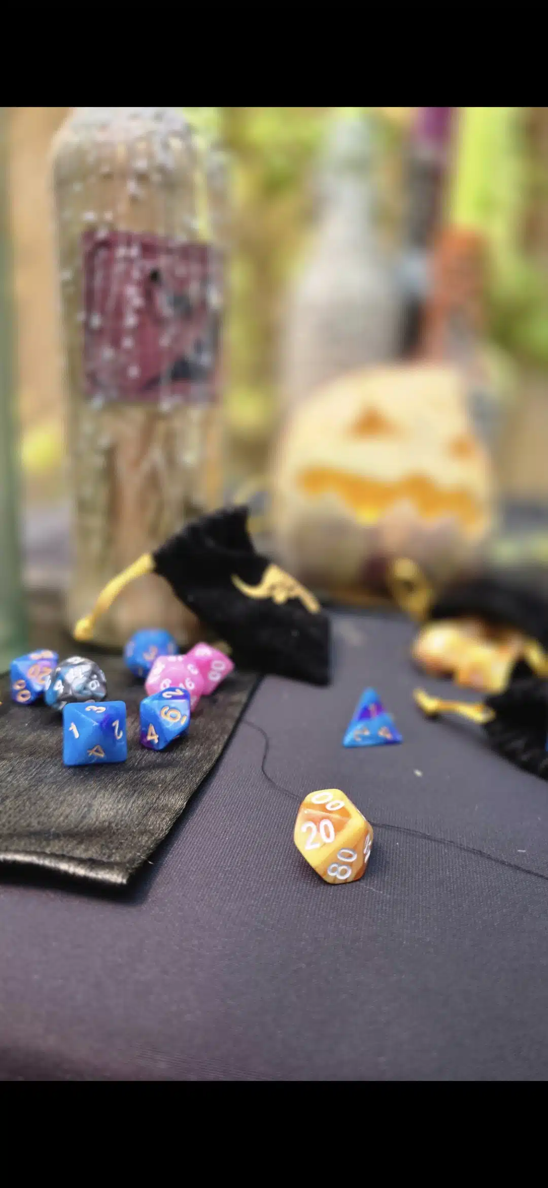 A set of colourful polyhedral dice, including an orange 20-sided die, are scattered on a black cloth near velvet bags, bottles, and a carved pumpkin in the background.