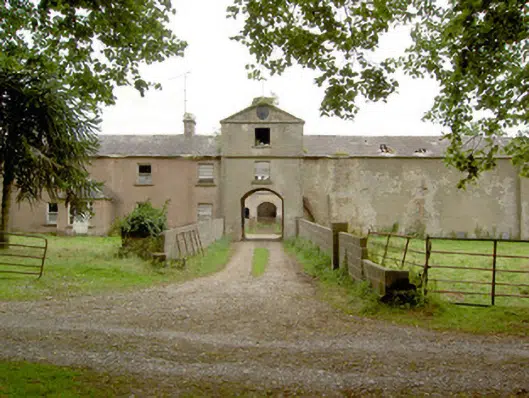 A gravel path leads to an old, weathered building with an arched entrance, surrounded by greenery and trees, with a metal gate and patchy grass on either side.