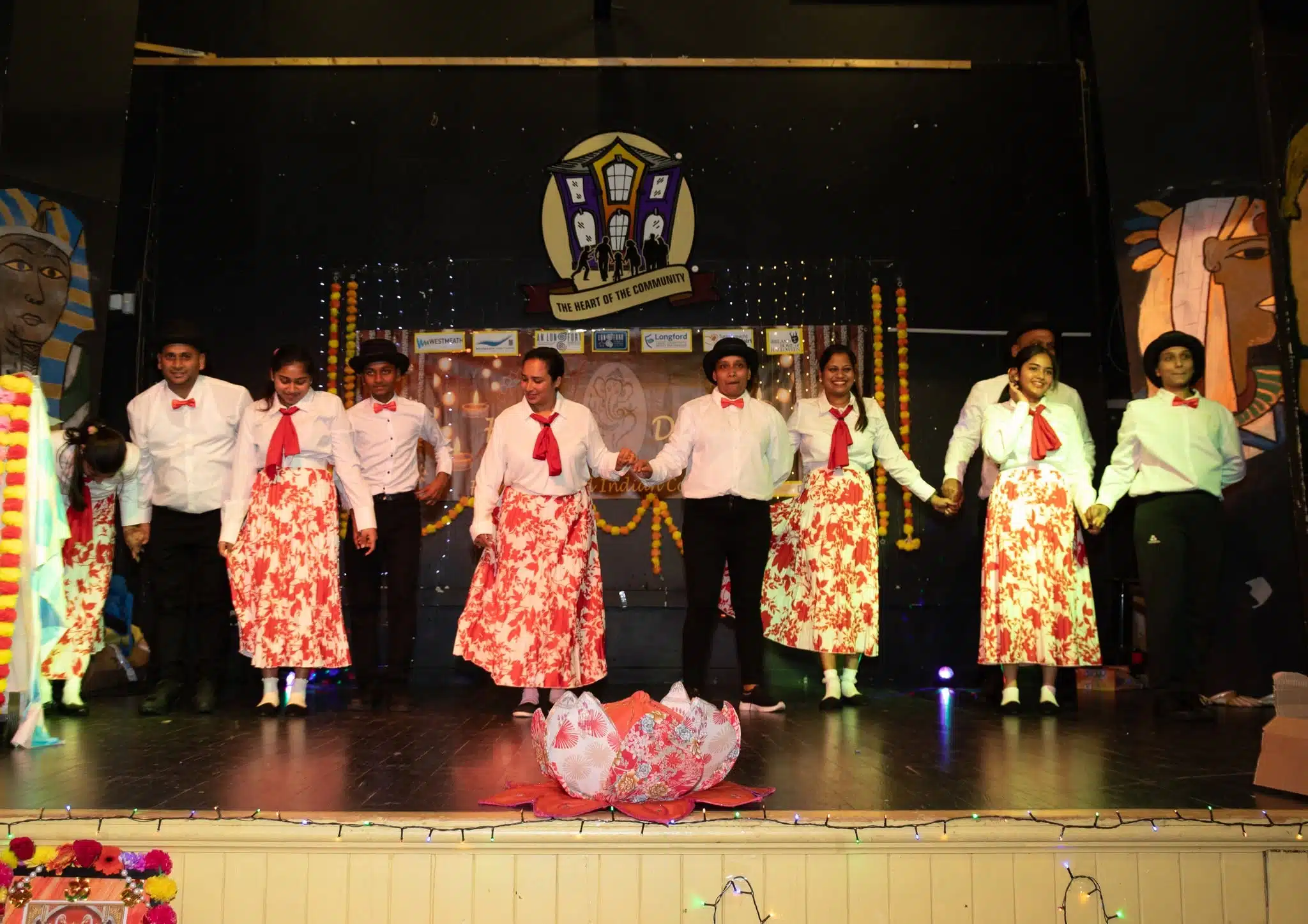 A group of performers in matching red and white costumes hold hands on stage, smiling after a performance, with festive decorations and props around them.