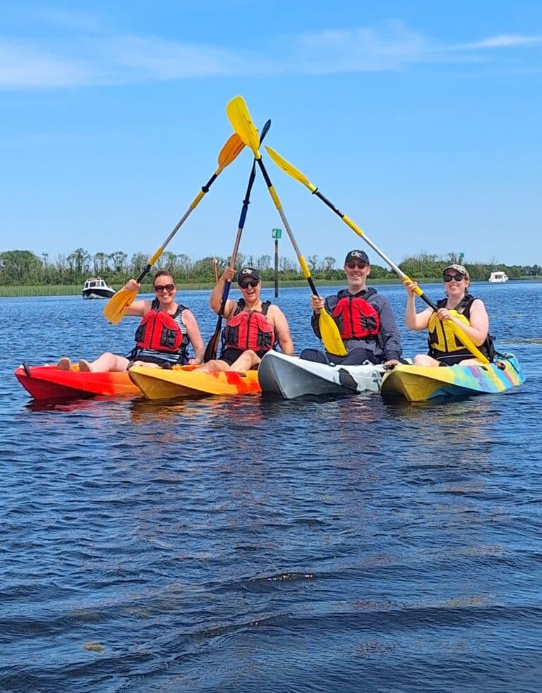 Four people in kayaks, wearing life jackets, raise their paddles together to form an X over calm water, with trees and boats visible in the background under a clear blue sky.