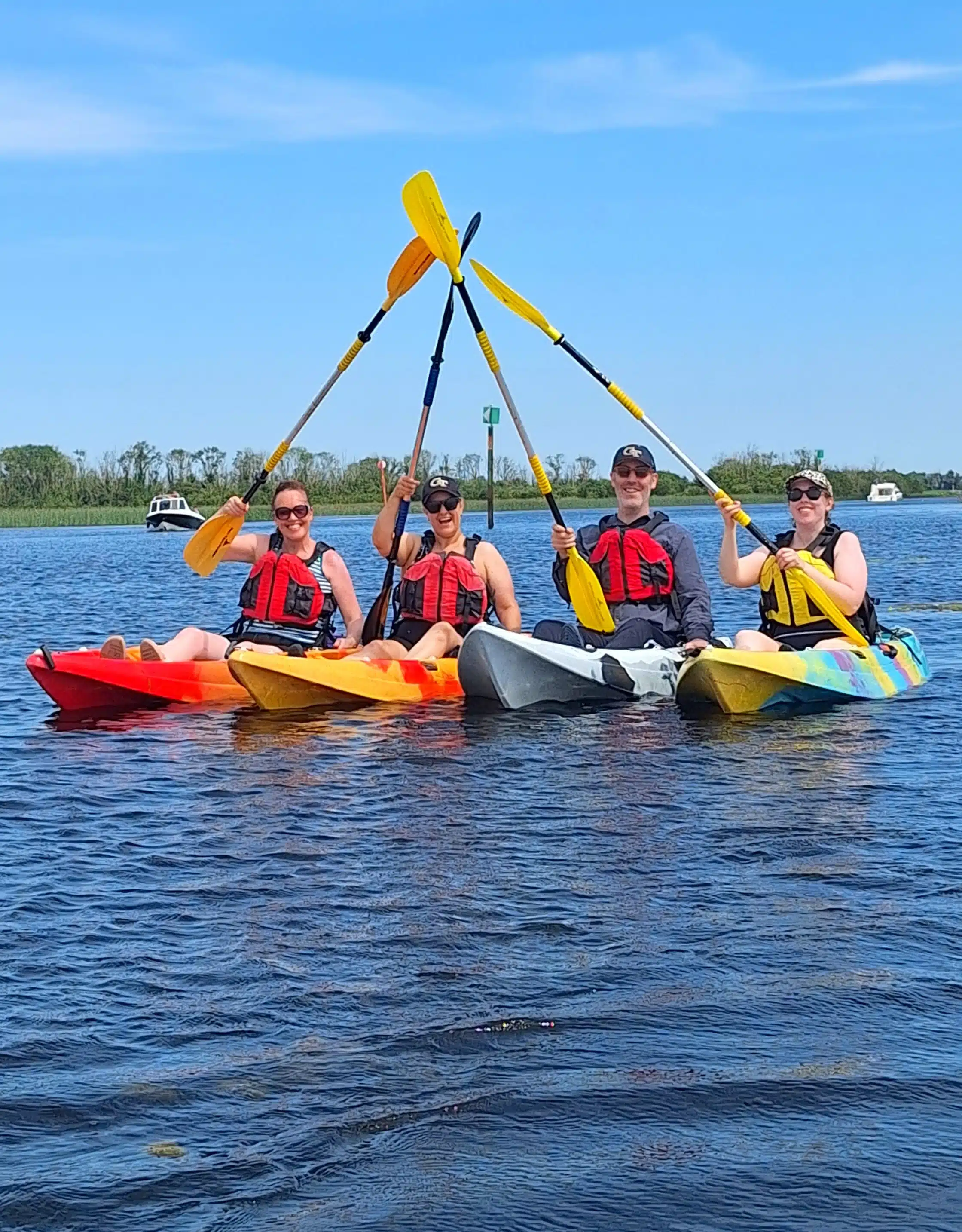 Four people in kayaks, wearing life jackets, raise their paddles together to form an X over calm water, with trees and boats visible in the background under a clear blue sky.
