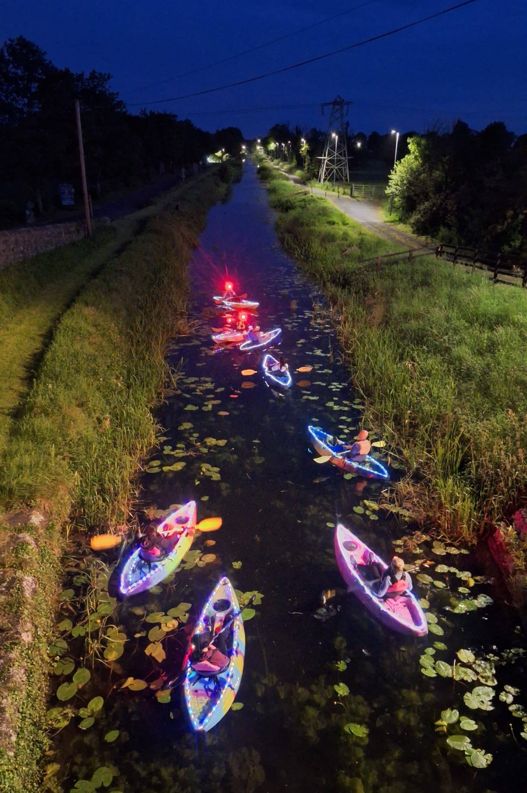 A group of people kayaking at night in a narrow canal, their kayaks decorated with colourful LED lights, surrounded by lily pads and grassy banks, with streetlights illuminating the path nearby.