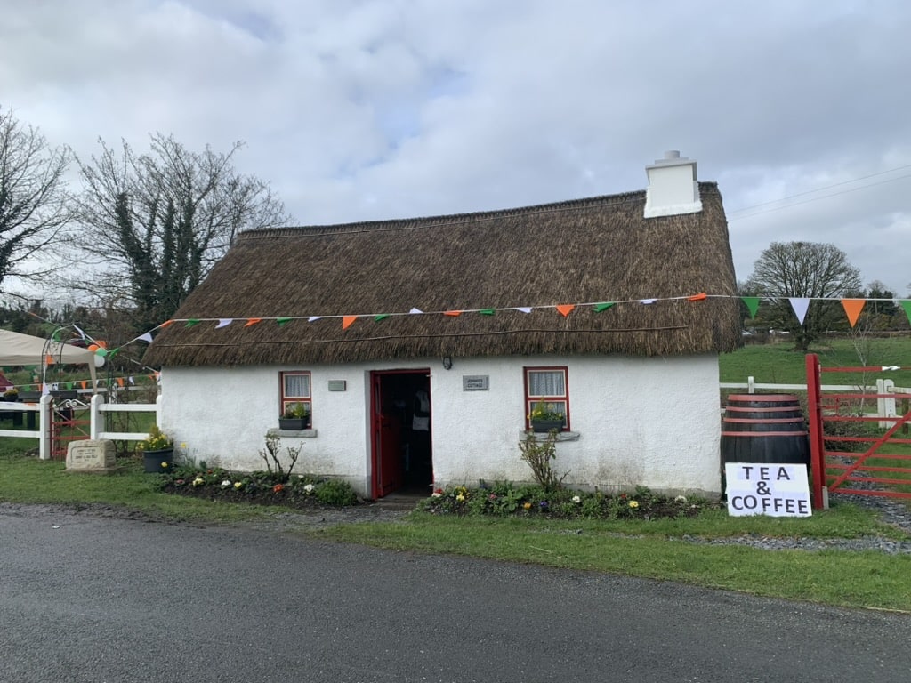 A small white cottage with a thatched roof, red door, festive bunting, flowers, and a sign outside reading "Tea & Coffee." The setting is rural with trees and green fields in the background.