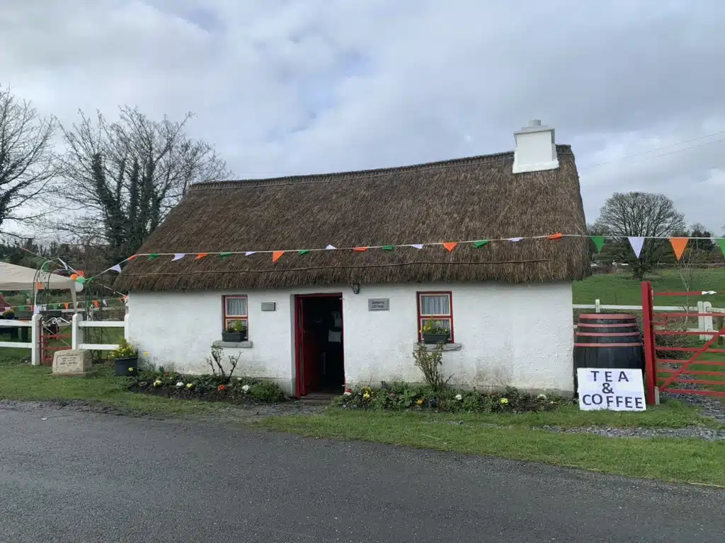 A small white cottage with a thatched roof, red door, festive bunting, flowers, and a sign outside reading "Tea & Coffee." The setting is rural with trees and green fields in the background.
