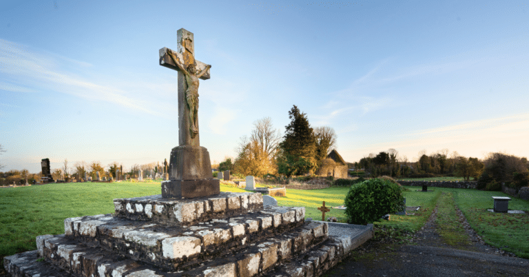 Large stone crucifix on a stepped base in a peaceful, grassy cemetery at sunrise, with headstones, trees, and a small building visible in the background.