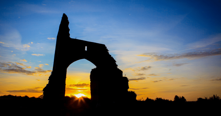 Silhouette of a ruined stone archway against a colourful sunset sky, with the sun low on the horizon and some clouds scattered above.