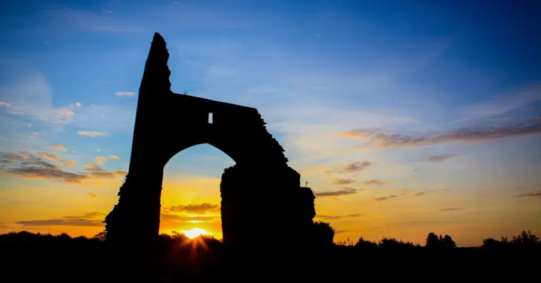 Silhouette of a ruined stone archway against a colourful sunset sky, with the sun low on the horizon and some clouds scattered above.