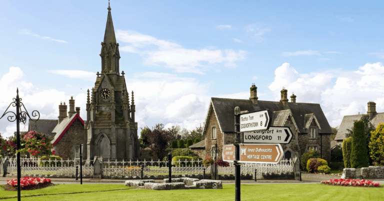 A stone church with a clock tower is surrounded by old buildings, green grass, and flower beds. Directional road signs stand in the foreground under a bright blue sky with scattered clouds.