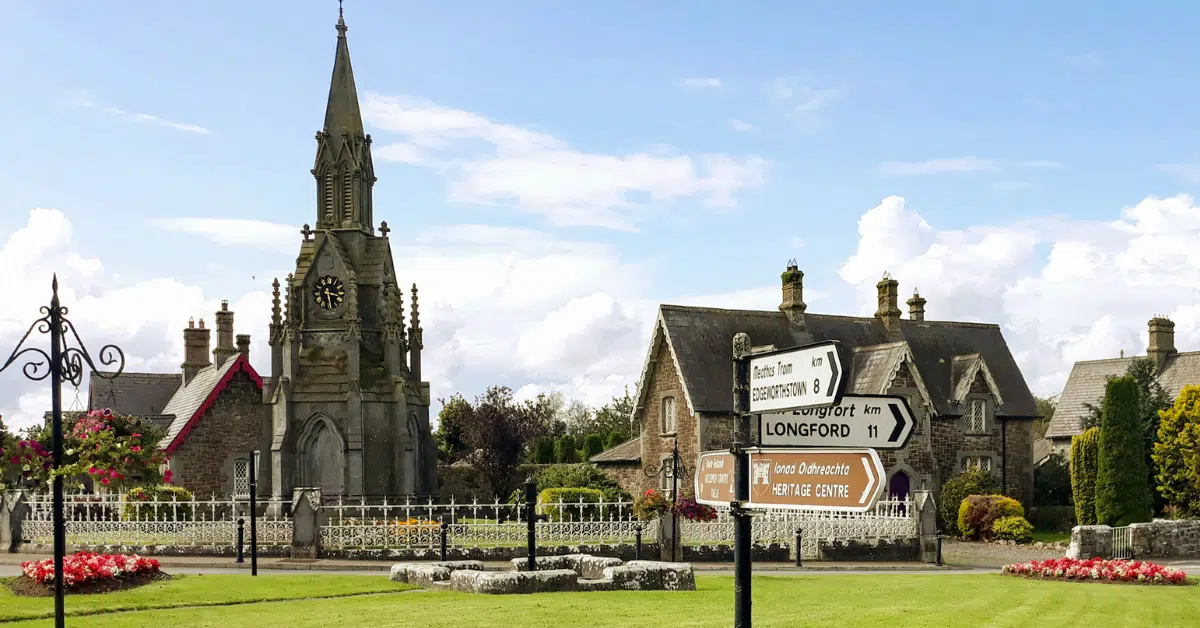 A stone church with a clock tower is surrounded by old buildings, green grass, and flower beds. Directional road signs stand in the foreground under a bright blue sky with scattered clouds.