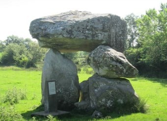 Large flat stones form a prehistoric dolmen in a grassy field with trees in the background. A plaque stands near the stones, indicating this site’s historical significance.