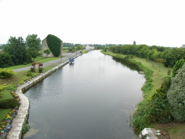 A calm canal runs between grassy banks with a small road and boat on the left. Flowerbeds and green shrubs line the water, and a few houses and trees are visible in the background under a cloudy sky.