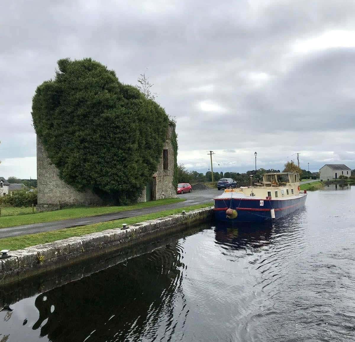 A canal boat moves past an old stone building heavily covered in green ivy, with cloudy skies above and a few cars parked along the narrow road beside the water.