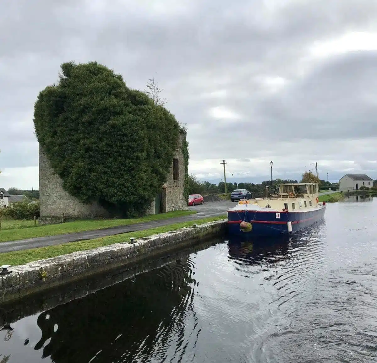 A canal boat moves past an old stone building heavily covered in green ivy, with cloudy skies above and a few cars parked along the narrow road beside the water.