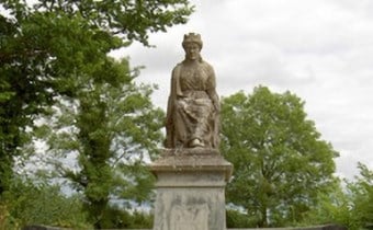 A stone statue of a seated crowned figure is positioned on a plinth outdoors, surrounded by green trees and under a cloudy sky.