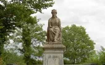 A stone statue of a seated crowned figure is positioned on a plinth outdoors, surrounded by green trees and under a cloudy sky.