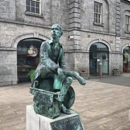 Bronze statue of a seated man with papers on his lap, positioned in front of a grey stone building with arched windows and doors.