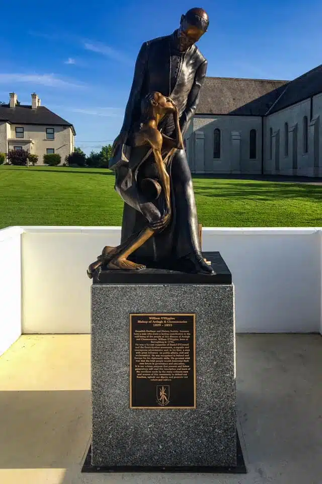 A bronze statue of a man holding a weak, emaciated person on a stone plinth with a plaque, set outdoors near grass and buildings under a blue sky.