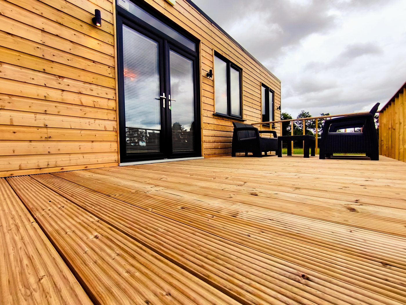 A modern wooden decking with black-framed glass doors, two black chairs, and a wooden balustrade, set against a cloudy sky.