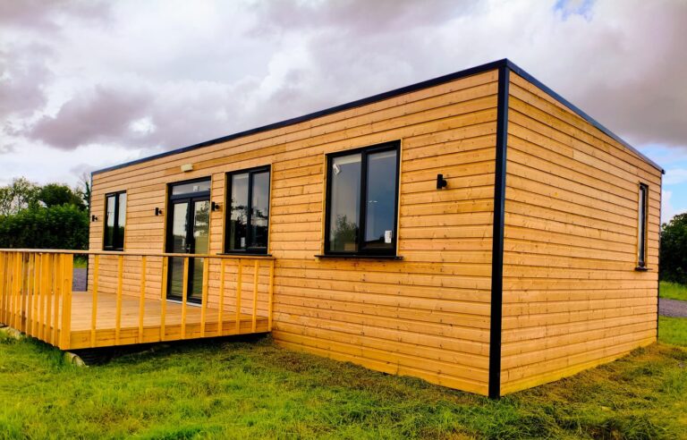 A modern tiny house with wooden exterior cladding, large windows, and a small front veranda sits on green grass under a cloudy sky.