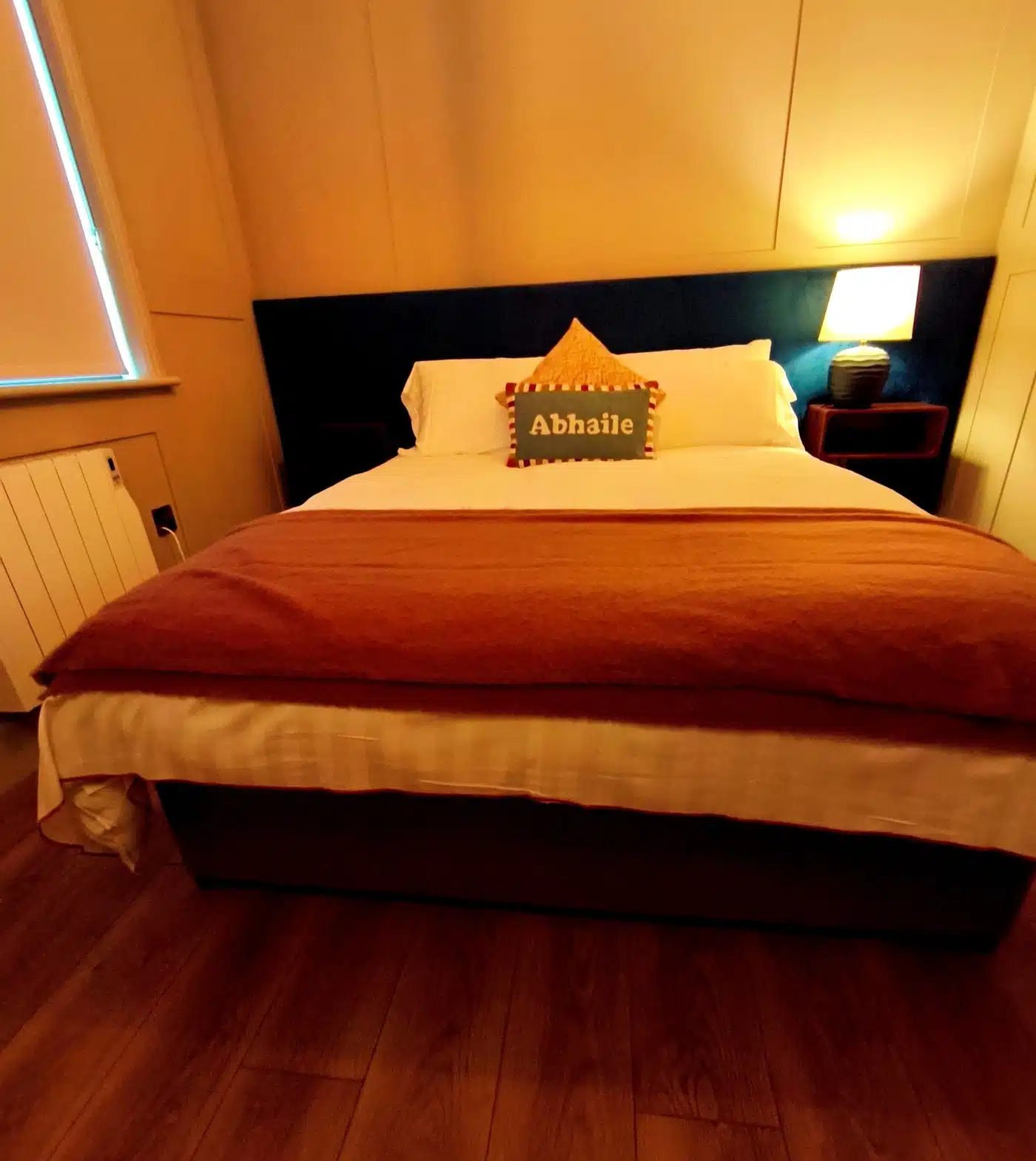 A cosy bedroom with a double bed, white and rust-coloured bedding, a pillow labelled "Abhaile," a lit bedside lamp, wooden floor, and a window with the blinds partially closed.