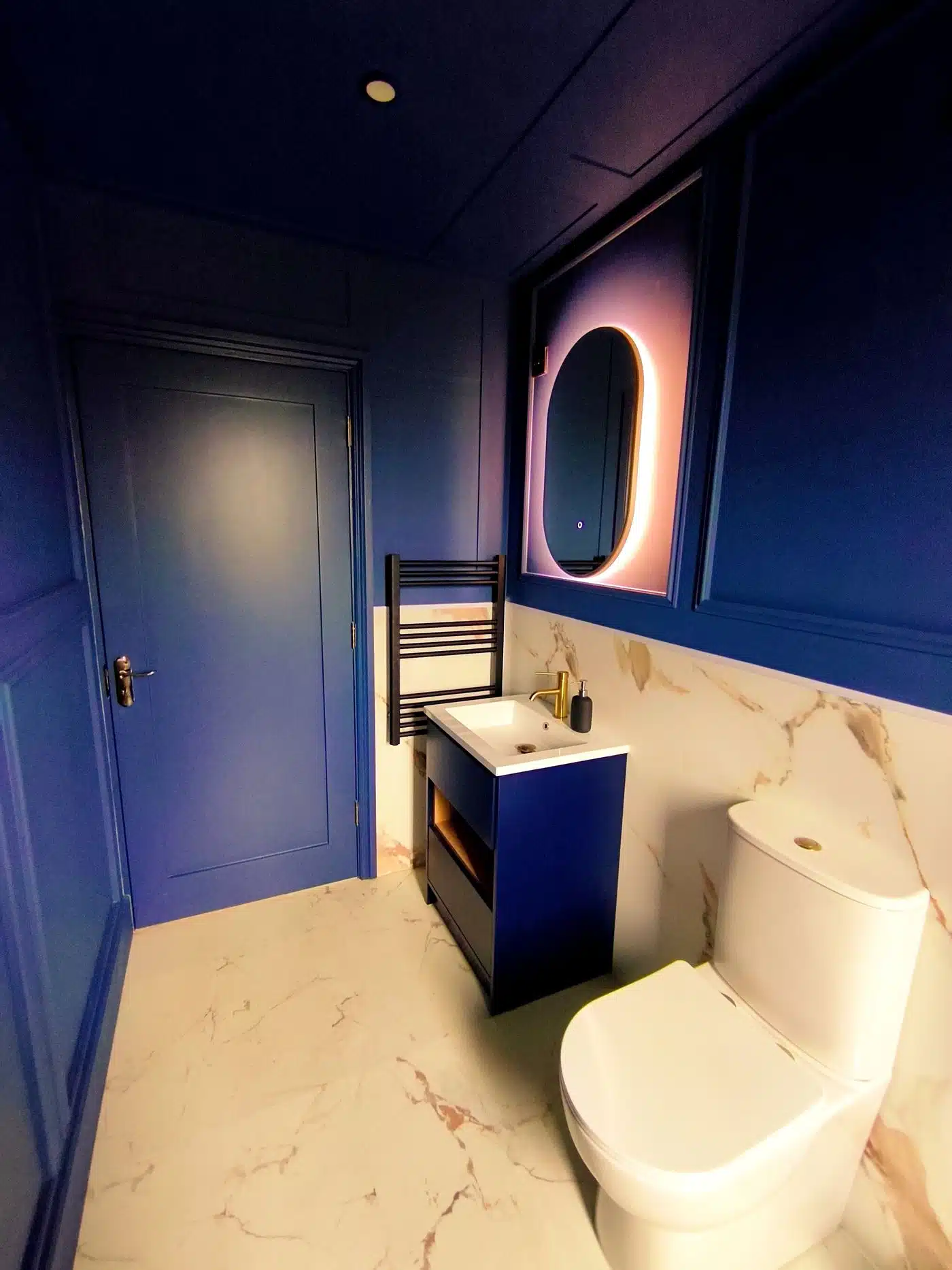 A modern bathroom with blue walls, a round LED-lit mirror above a basin, a white toilet, marble-patterned flooring, and a heated towel rail beside the blue door.