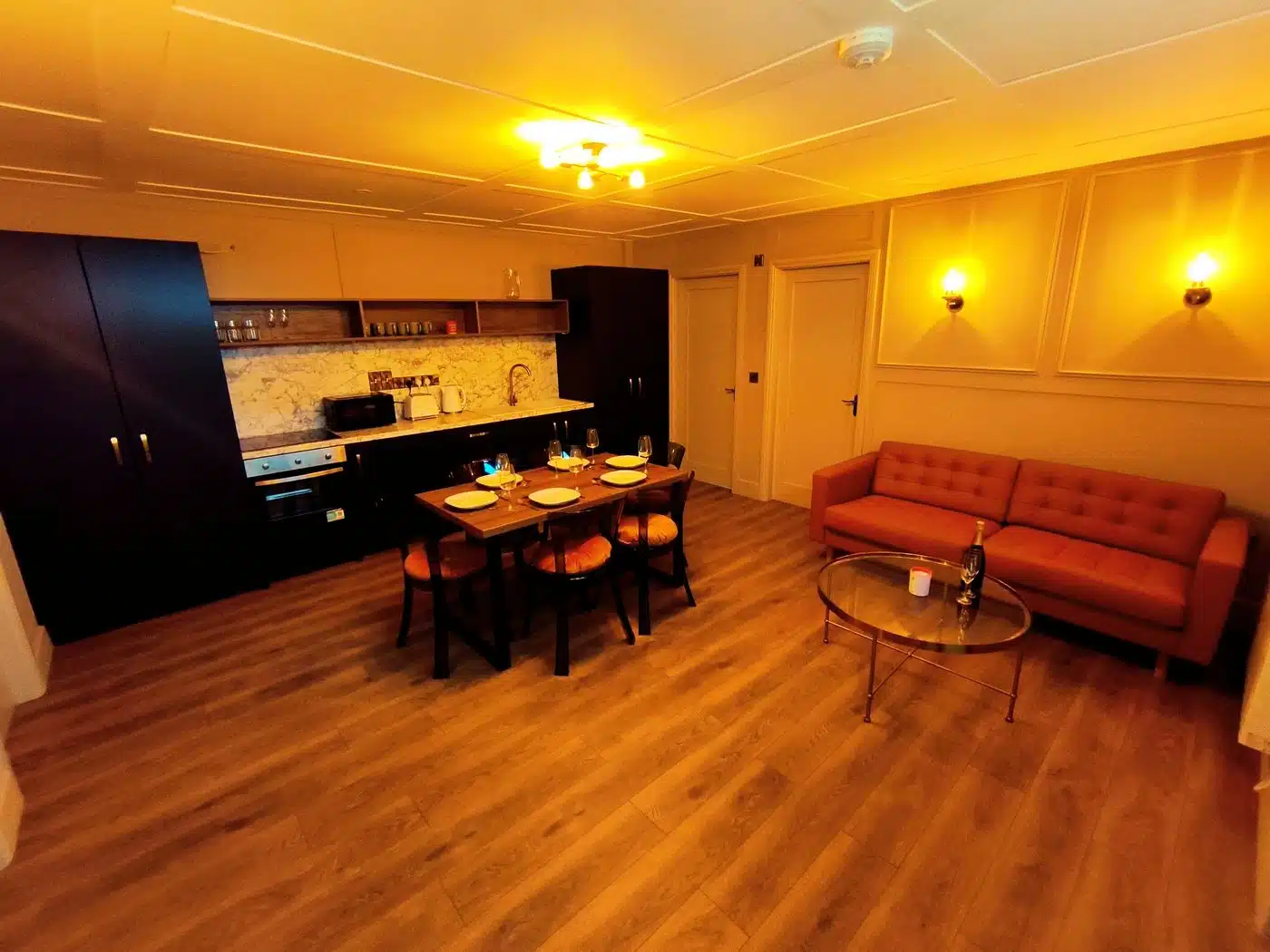 A cosy kitchen and living area with wooden floors, orange sofa, glass coffee table, dining table set for four, and dark cupboards against a wall with marble splashback. Warm yellow lighting fills the room.