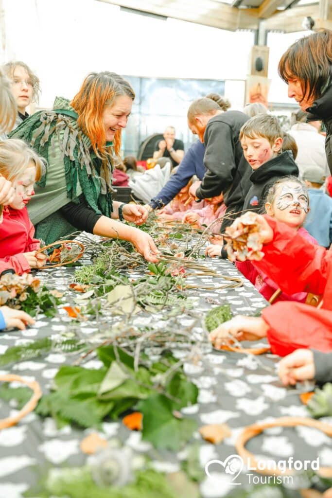 Adults and children gather round a table with leaves, twigs, and craft materials. Some kids have face paint, and everyone is smiling, enjoying an arts and crafts activity indoors that celebrates Sustainable Tourism Longford. The Longford Tourism logo is visible.