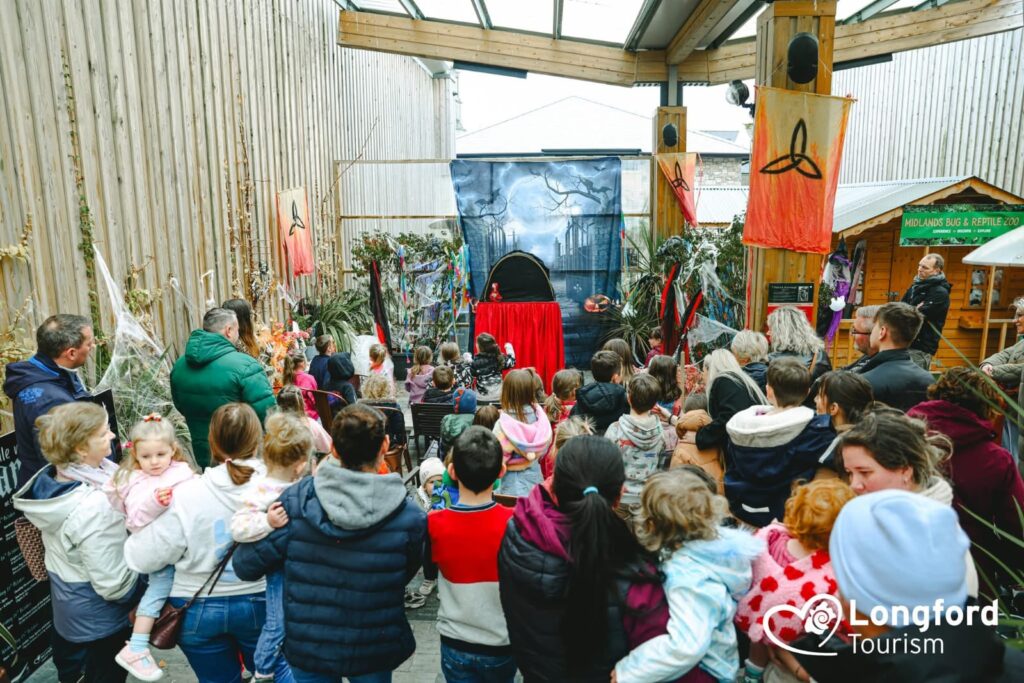 A crowd of adults and children watch a puppet show inside a covered outdoor area decorated with flags and plants, celebrating the spirit of Sustainable Tourism Longford. The Longford Tourism logo is visible in the bottom right corner.