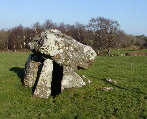 Large ancient stone structure stands on a grassy field, composed of upright stones supporting a massive flat stone. Leafless trees and blue sky appear in the background.