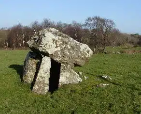 Large ancient stone structure stands on a grassy field, composed of upright stones supporting a massive flat stone. Leafless trees and blue sky appear in the background.