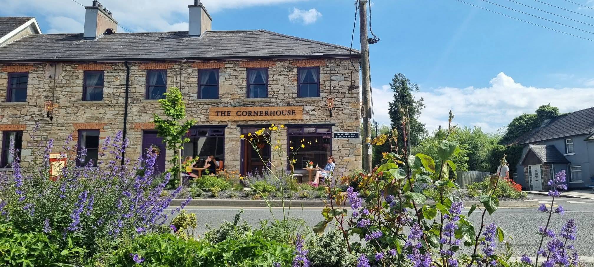 A stone building labelled "The Cornerhouse" sits behind a garden filled with purple flowers on a sunny day. People are seated outside at tables, and a road runs in front of the scene.