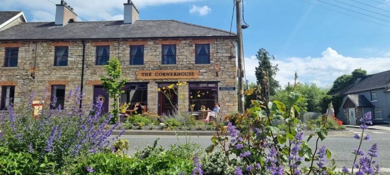 A stone building labelled "The Cornerhouse" sits across the street, with people at outdoor tables and purple flowers in the foreground under a bright blue sky.