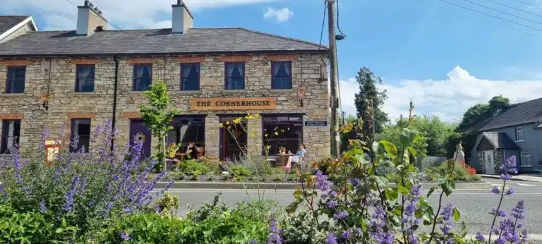 A stone building labelled "The Cornerhouse" sits across the street, with people at outdoor tables and purple flowers in the foreground under a bright blue sky.