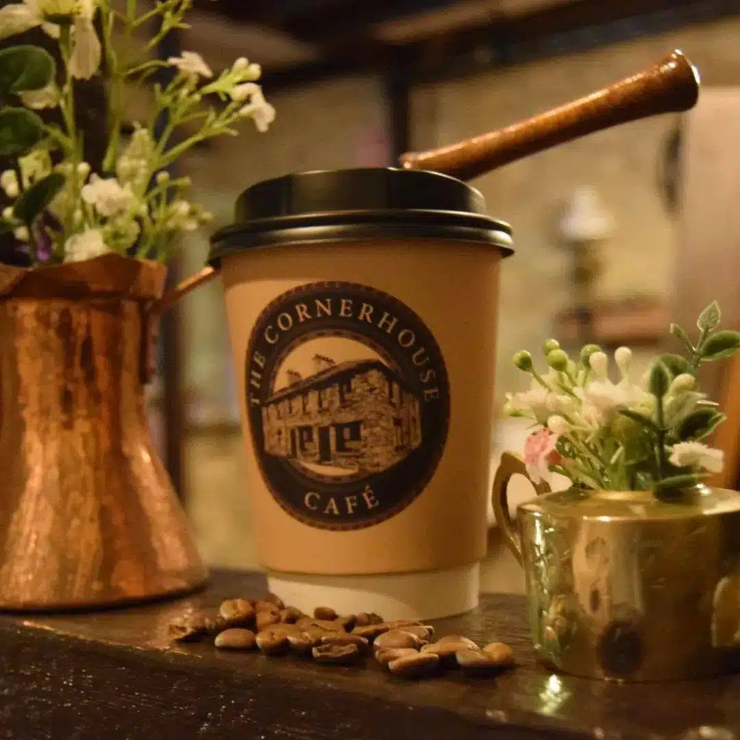 A coffee cup labelled "The Cornerhouse Café" sits on a wooden surface, surrounded by flowers in copper containers and scattered coffee beans.