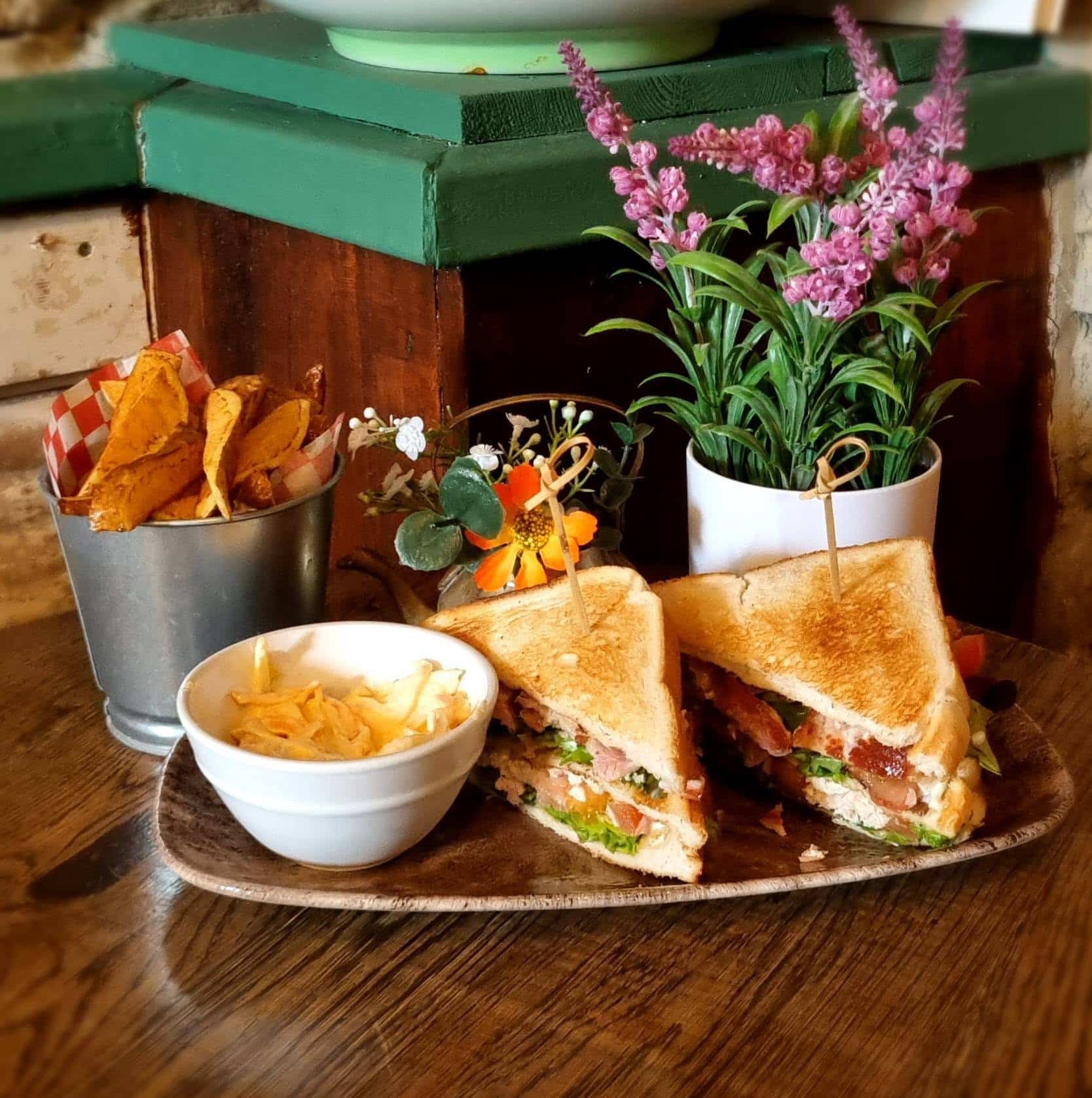 A plate with a sandwich cut in half, a small bowl of coleslaw, and a bucket of crisps. There are potted flowers in the background on a wooden table.