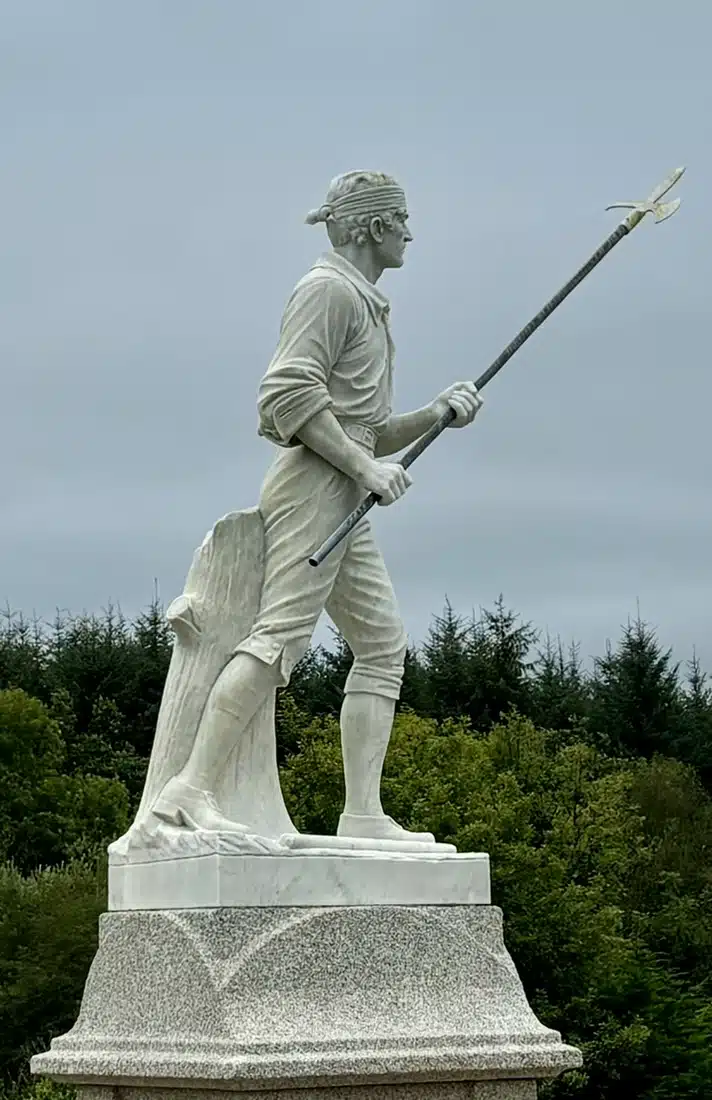 A white statue of a man in colonial attire holding a spear, standing on a granite plinth with trees and a cloudy sky in the background.