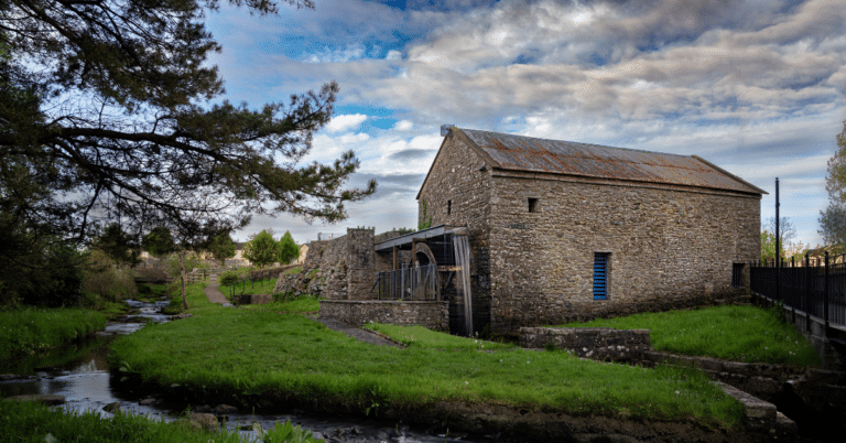 A stone watermill with a rusted roof stands beside a calm stream, surrounded by green grass and trees, under a partly cloudy sky.