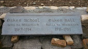 A stone plaque reads: "Esker School Scoil na hEasrach 1869-1914" and "Esker Hall Halla na hEasrach 1915-1981," resting on stones, with gravel and rocks in the background.