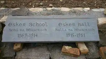 A stone plaque reads: "Esker School Scoil na hEasrach 1869-1914" and "Esker Hall Halla na hEasrach 1915-1981," resting on stones, with gravel and rocks in the background.