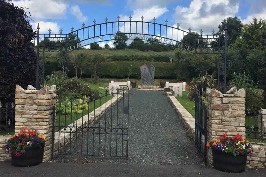 An open iron gate reading "Garden of Remembrance" leads to a gravel path flanked by stone walls, benches, and two stone memorials, with colourful flowers in planters and greenery in the background.
