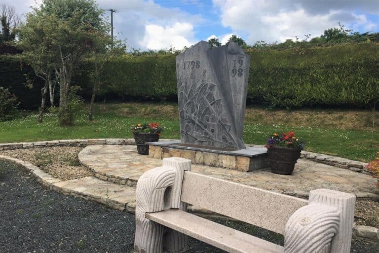 A stone bench faces a grey memorial inscribed with "1798" and "1998," set in a circular stone area with two flower pots and surrounded by grass, trees, and a hedge under a partly cloudy sky.